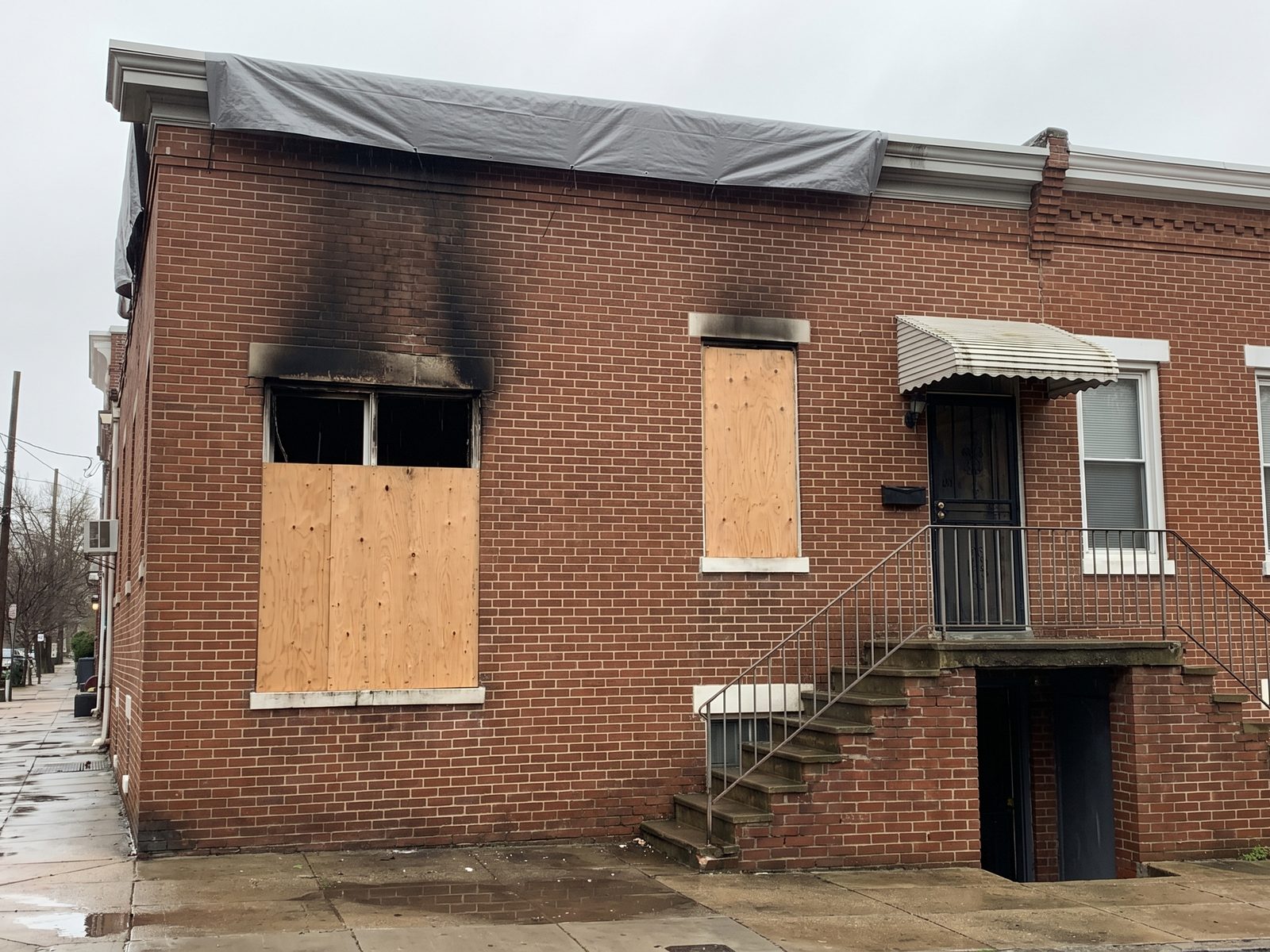 Boarded window and roof tarp after a row-house fire Soot marks above a boarded window on an attached brick row house with wet pavement and a tarp on the roofline above.