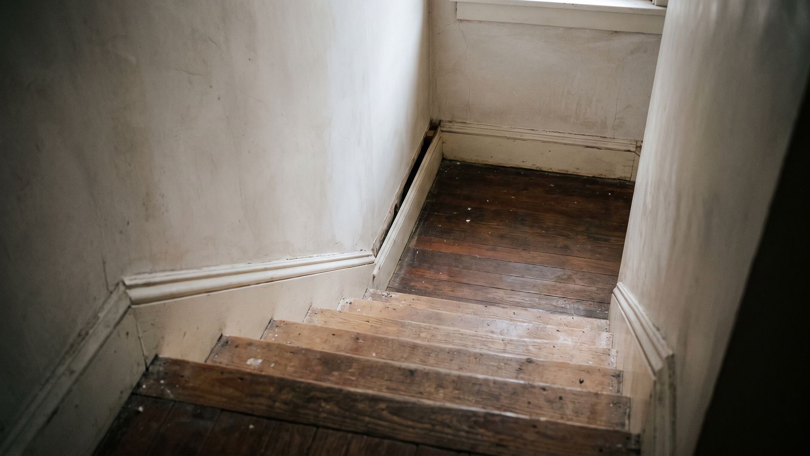 A stair landing in a row house, bare wood treads, one length of baseboard standing loose from the plaster wall.