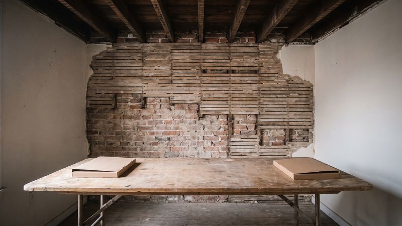 Two closed folders at opposite ends of a bare wooden table in a row house room with plaster cut back to the lath.