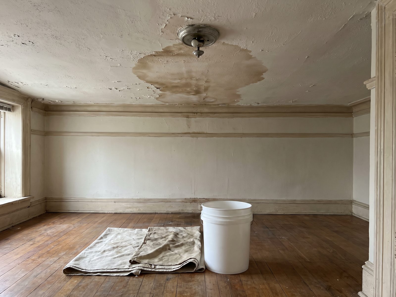 Ceiling leak into the unit below Wet patch spreading across a painted plaster ceiling from a light fixture with a drop cloth and bucket below.