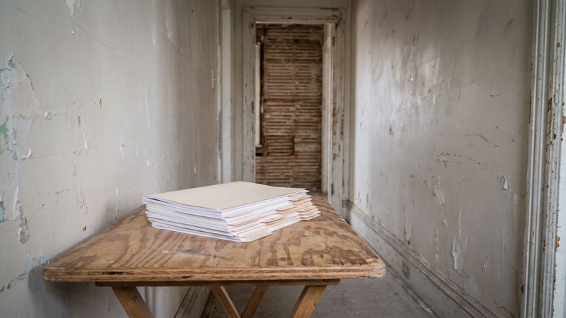 A tabbed document set stacked on a plywood folding table in a stripped rowhouse-style corridor