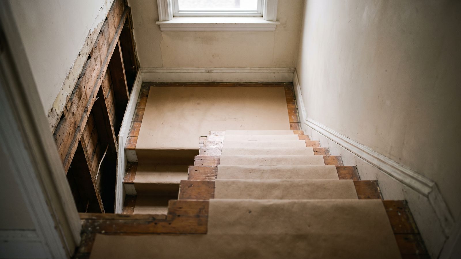Stair treads covered edge to edge with protective paper beside a wall opened to the lath and studs
