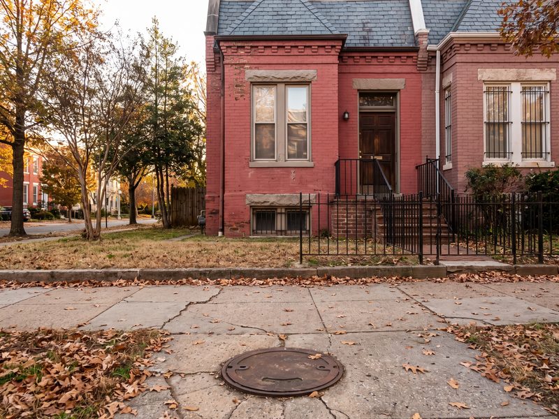 A round cast-iron utility lid set into the public sidewalk in front of a brick row house.