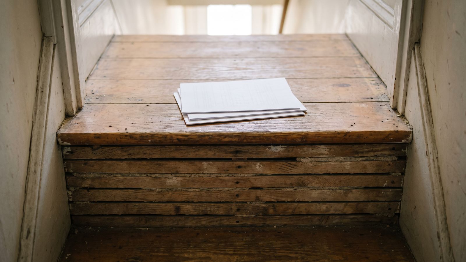A small stack of unmarked pages on a bare stair tread in a row-house stairwell stripped back to lath
