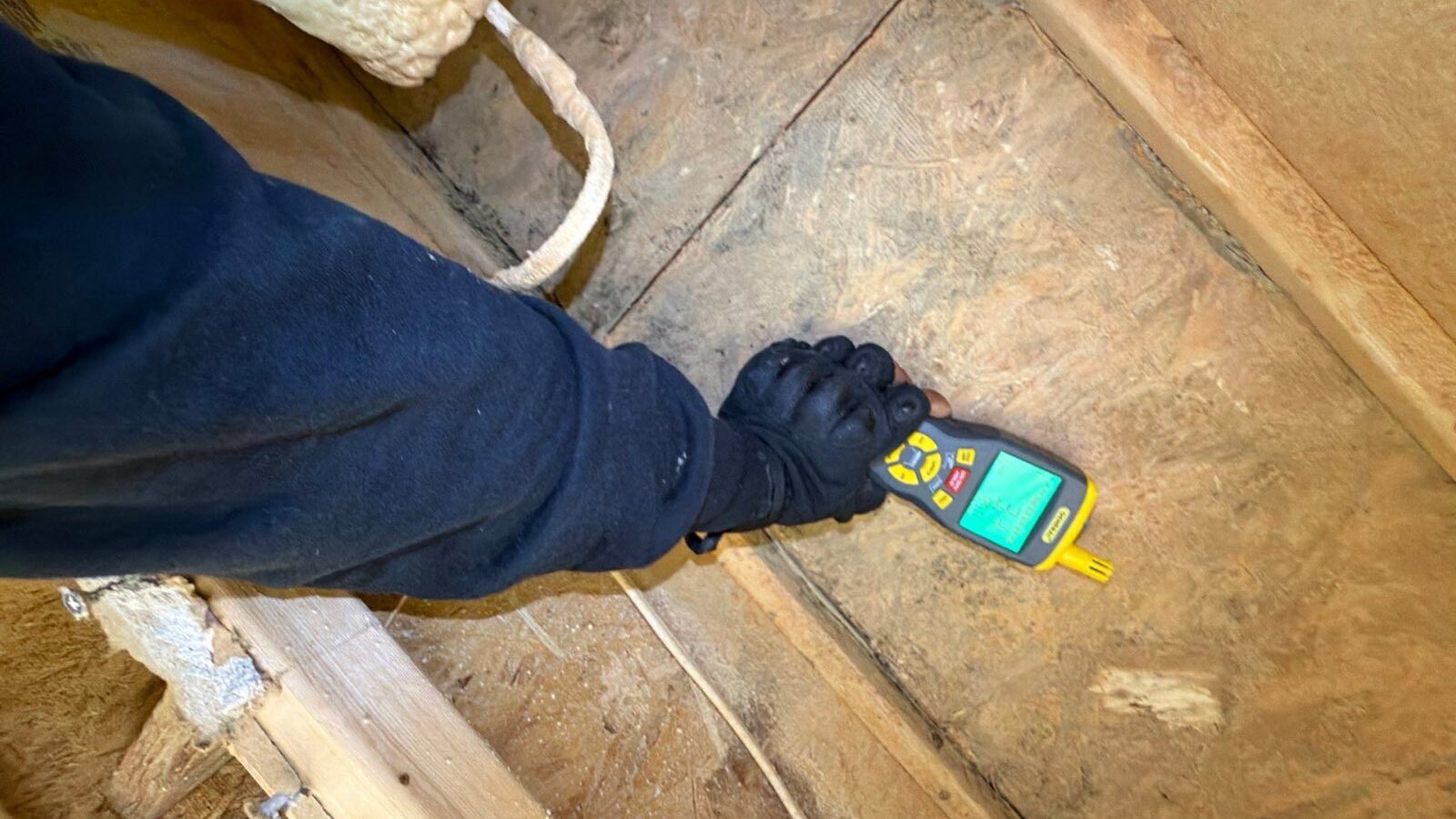 A gloved hand presses a handheld moisture meter against bare subfloor boards between floor joists