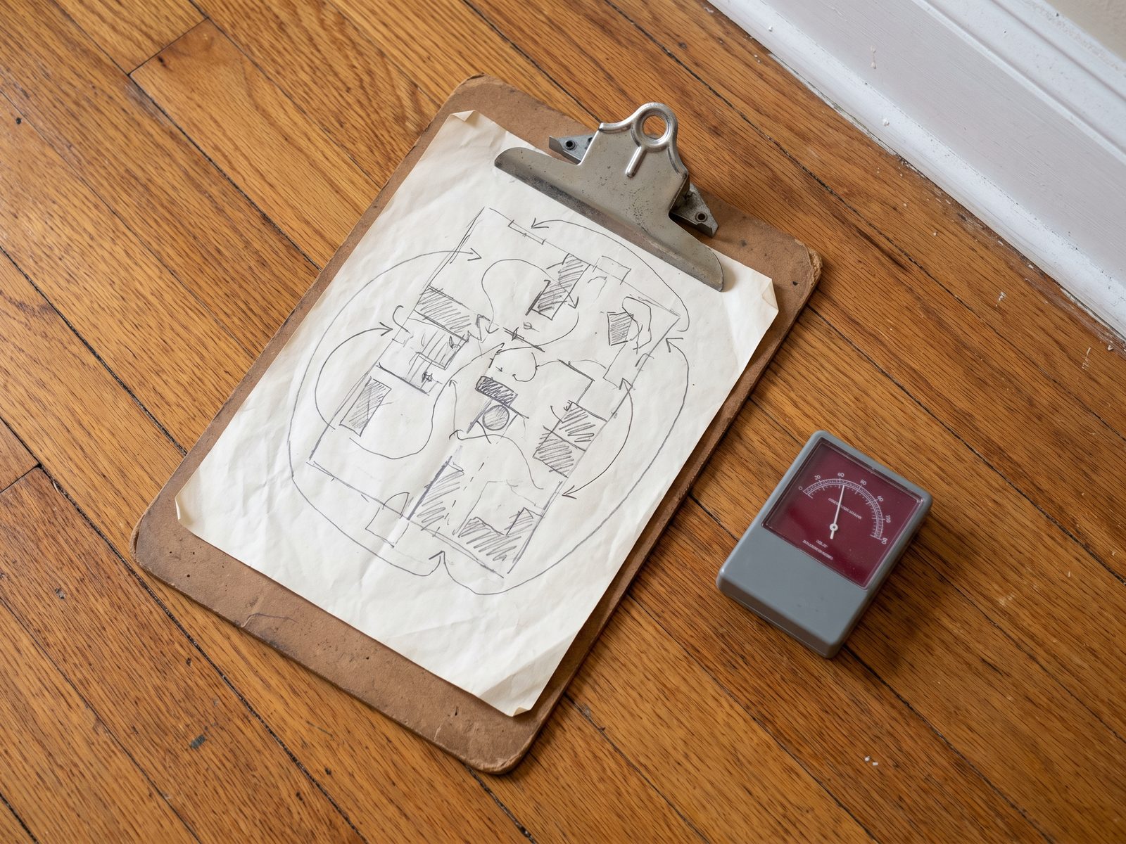 Drying plan and hygrometer on a hardwood floor Hand-drawn drying plan on a clipboard next to a dial hygrometer resting on a hardwood floor.