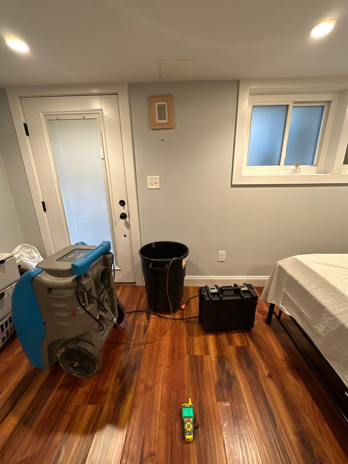 Blue dehumidifier, black bin, equipment case and moisture meter on a dark-stained wood floor in a bedroom in Washington, DC, July 2025