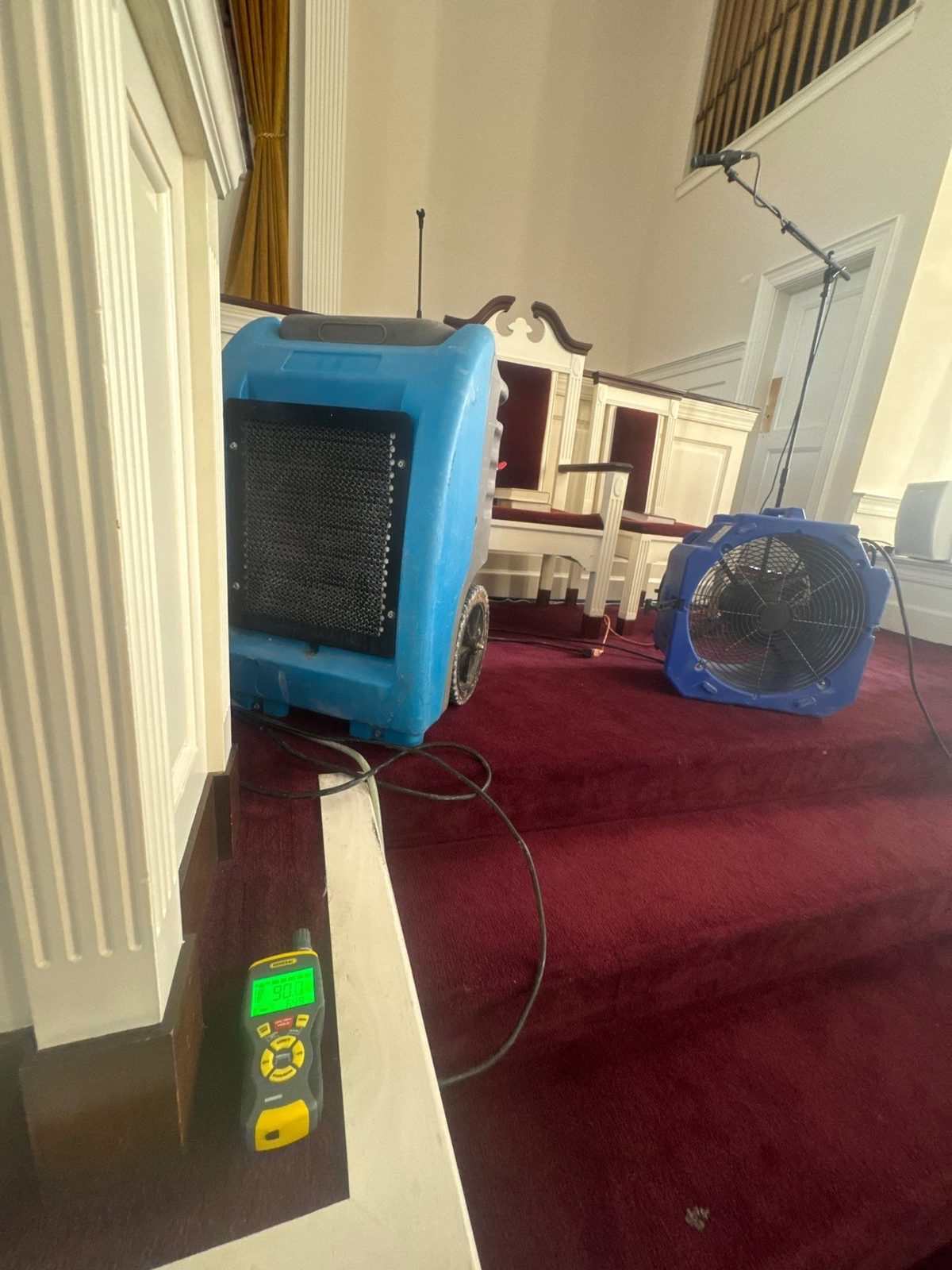 Blue dehumidifier and air mover on carpeted steps beside a moisture meter in a sanctuary in Washington, DC, March 2025