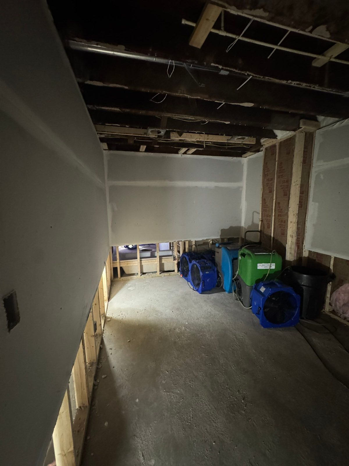 Gutted room with drywall removed to the studs and blue and green dehumidifiers and air movers on a concrete floor in a rowhouse in Washington, DC, September 2026